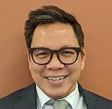 close-up of Tony Flores wearing a light-coloured shirt, tie, and dark blazer, smiling directly at camera