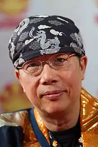 Profile of Toshio Maeda looking at the camera wearing glasses and dark blue patterned head covering.