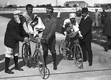 Two men with flowers in their hands, sitting on bicycles being held by other men, with the Eiffel tower seen in the background.