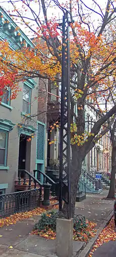 A black latticework metal needle-shaped tower in a city street with the leaves in autumn color