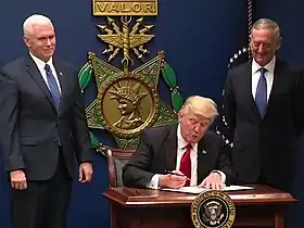 Donald Trump signing the order in front of a large replica of a USAF Medal of Honor, with Mike Pence and James Mattis