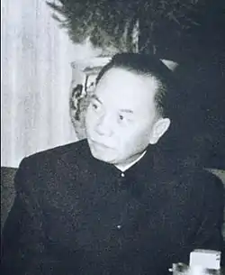 A balding man looking to the left, dressed in a dark jascket buttoned to the neck