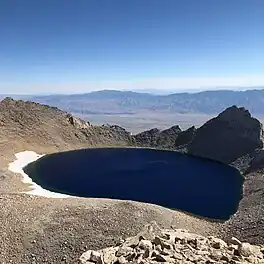 Tulainyo Lake with nearly black water, rimmed with snow, seen from above looking out at the lake and Owens Valley beyond the ridge to its east