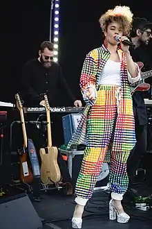 Pale skinned black female wearing platform shoes and a multi coloured patchwork trouser suit singing on stage with a background of racked spare guitars, one guitarist and a keyboard player