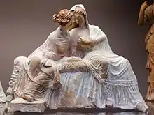 Terracotta statue of two women, wearing long dresses, seated and leaning on each other.