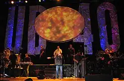 The members of UB40 are performing on a stage, most of whom are wearing black.  A light focuses on the lead singer in the middle of the image, singing with a microphone in hand.  The background of the stage has "UB" and "40" spelled in large letters with an oval shape in the middle.