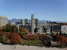 Photograph of a clock tower, standing in a park framed in autumn coloured trees. Beyond the tower are six high-rise grey buildings and behind those are mountain peaks.