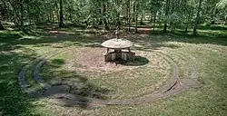 A recent photograph of the monument shows a concrete model of the spaceship in a clearing. There are concrete paths beneath and around the model in the clearing. In the background, a forest is visible with two trails.