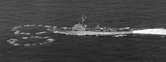 A destroyer moving to the left; on the water just ahead of her are two rings of small circular waves, one to either side of her path. The rings nearly meet directly ahead of her and are each about half her length in diameter.