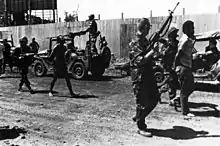 Two men in civilian clothes with their hands on their backs walk surrounded by three armed men in uniform. Military jeeps are seen in a second plane.