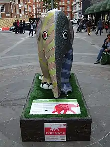 A statue of an elephant is brightly painted in two halves: the left half in lines of yellow and dots of orange; and the right half in overlapping scales in gray, and bands of light and dark blue. The statue sits on a base with the appearance of black rock, an information plate at the front describing the artwork. It is situated in the middle of the pedestrianised road next to Harrods, and people are sitting and walking around in the background.