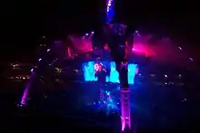 At night in a large outdoor stadium filled with people, a structure with four legs curving upward and meeting in a central spire is bathed in indigo and violet light, as a large circular video screen below the spire but above a stage shows a dark-clothed man with arm raised singing into an illuminated circular microphone.