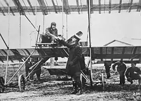 Pilot seated in open-framed  biplane, shaking hands with a man in military uniform who wears a peaked cap