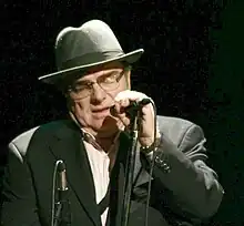 Man wearing a grey hat and grey jacket and glasses holding a microphone near his face.