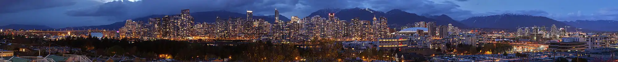 High-resolution panorama of a large, brightly lit skyline at night. A mountain range lies in the background, and a bridge is visible on the left-hand side of the panorama.