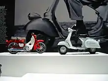 A 1960s Italian scooter and a 1960s single cylinder Japanese motorcycle in front of a giant image on the wall of the front wheel and fender of a 1960s Italian scooter.