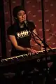 A woman plays keyboard and sings in front of a red patterned background.