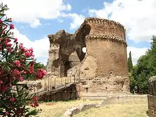 Ruined cylindrical brick building exterior with a large section missing showing the hemispherical domed interior with what had been four round windows in the sides of the dome and no oculus at the top