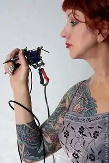 Vyvyn Lazonga holding a tattoo machine and displaying her tattooed inner right arm.