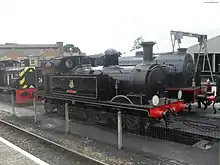 A steam locomotive in a maintenance depot. Beneath it is an inspection pit. To the left is a Diesel shunting locomotive; to the right is another steam locomotive and a crane.