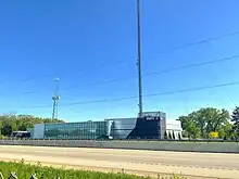 A modern two-story building with WMTV and Madison CW's logos, along with the station's transmitter behind the building along a freeway.