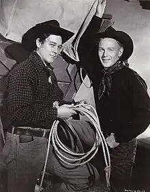 Black and white photograph showing two men standing. They are dressed in cowboy garb: jeans, kerchiefs around their necks, and cowboy hats. The man on the left is holding a lasso near his waist. A section of what appears to be a covered wagon is visible behind them.