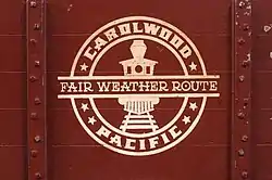 A circular, white-colored logo consisting of a forward-facing steam locomotive in the center, the text "Carolwood Pacific" around the edge, and the text "Fair Weather Route" across the middle. The logo is painted on the side of a miniature, reddish-brown-colored freight car.