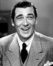 Black and white portrait of a smiling white man with dark hair. He is wearing a three-piece suite with a dark tie and a white handkerchief in his breast pocket.