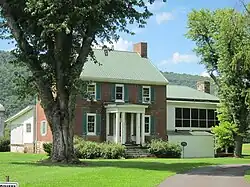 An image of two-story brick house with a green roof, a large front porch, and a tree in the foreground.