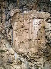 The photograph shows a sculpted rock: a bas-relief appears in grey-beige on a blue-gray rock stained with dark grey streaks. The Hittite king is on the left. He is bearded and holding a branch with two grapes. Facing him, the god has lost his head and his feet because of the ravages of time