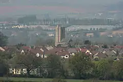 Roofs of several houses with the square tower of the church prominent amongst them. In the background are fields and hills.