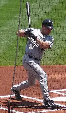 A man in a gray baseball uniform and black batting helmet stands in the batter's box twisting from the waist while holding a baseball bat