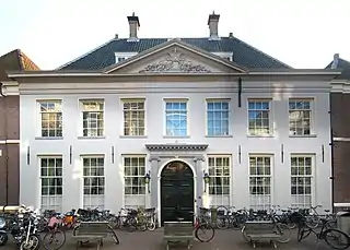 This is a white building, standing two stories tall and with a gabled roof. It has seven windows from its left to right side. The door is large with an arched top, painted black. The building has a pediment at the roof level which is roughly one-third the width of the whole building. Over the entry door, it reads in script "West Indisch Huis".