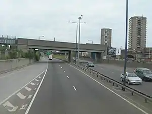 A dual carriageway crossed by a railway bridge in the foreground. Two high-rise blocks stand in the distance.