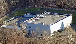 A large light-colored building with a flat roof seen from above, surrounded by bare trees.