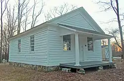 A white clapboard-sided one-story wooden building with a pointed roof and small front porch, seen from its right. Behind it are bare trees and a dusky sky