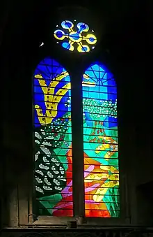 A stained-glass window in bright primary colours depicting a path in the countryside with trees and plants on either side