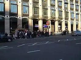 A large crowd of people walking on a grey sidewalk next to a black road where two vehicles are driving from the left to the right