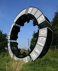 Modern sculpture using wheeled tubs from mineral mining attached to each other forming a riased circle ahainst a bright blue sky