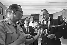 General Earle Wheeler holding up a model of a U.S. Army helicopter as Ambassador Vu Van Thai and President Lyndon B. Johnson look on.