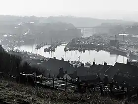 A black and white image looking upriver from Whitby Abbey. It shows the wide estuary harbour with many pleasure boats in the marina