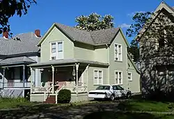 Photograph of a modest two-story wooden house with porch, closely flanked by other houses