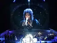 Houston performing on stage. Behind her is a screen where she is shown wearing a white outfit with a black overcoat and performing.