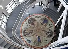 An aerial view of a mosaic depicting five people working, mining, farming -- all radiating out from the center like a flower. There are also tools of various trades like a hammer and a scythe surrounding the flower-like part.