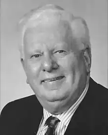 Black and white photo of a late middle-aged man wearing a dark-coloured suitjacket and tie, and white dress shirt with stripes