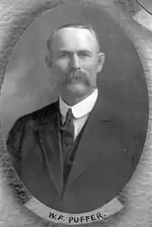 A torso shot of a middle-aged white man with receding dark hair and a large moustache, wearing a three piece suit