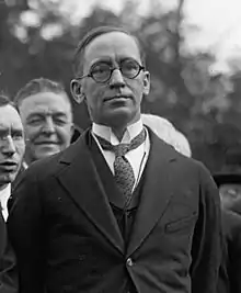 Black and white photograph of man with short, wispy hair wearing circular glasses, necktie, suit, and overcoat