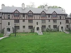 A three-story grey Tudor Revival building with reddish beams stands before green lawn on an overcast day.