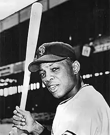 A right-handed batting baseball player with SF on his black cap stares towards the camera, with a baseball grandstand in the background