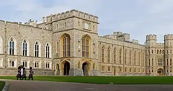 A photograph of a grey Gothic building stretching across the picture; the nearest parts have windows in a white stone, the furthest parts in a yellow stone. Three soldiers in grey uniforms and black hats are marching past the building.